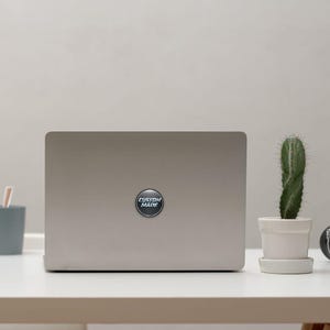 Puede incluir: Un port&aacute;til plateado con una pegatina circular que dice "CUSTOM MADE" en texto blanco. Un peque&ntilde;o cactus en una maceta blanca est&aacute; a la derecha del port&aacute;til. El port&aacute;til est&aacute; sobre un escritorio blanco.