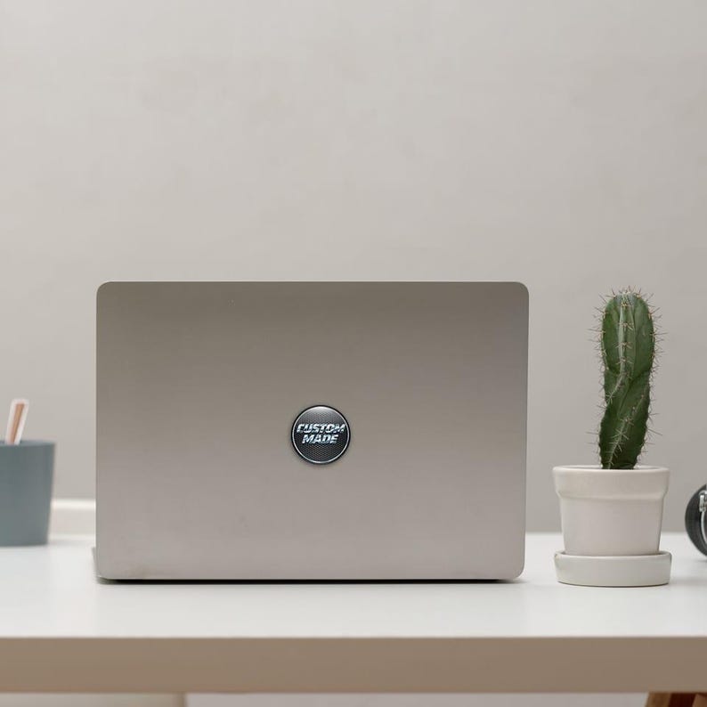 Puede incluir: Un port&aacute;til plateado con una pegatina circular que dice "CUSTOM MADE" en texto blanco. Un peque&ntilde;o cactus en una maceta blanca est&aacute; a la derecha del port&aacute;til. El port&aacute;til est&aacute; sobre un escritorio blanco.