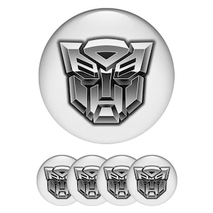 May include: A set of five white, round buttons featuring a metallic silver Autobot symbol. The buttons have a glossy finish and a 3D effect, with the Autobot logo prominently displayed on each.