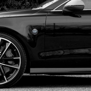 Puede incluir: Un coche negro con una llanta plateada y un espejo lateral. Un emblema cromado con las palabras "CUSTOM MADE" est&aacute; fijado a la carrocer&iacute;a del coche. La imagen es en blanco y negro.