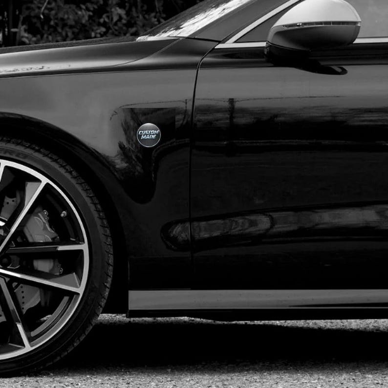 Puede incluir: Un coche negro con una llanta plateada y un espejo lateral. Un emblema cromado con las palabras "CUSTOM MADE" est&aacute; fijado a la carrocer&iacute;a del coche. La imagen es en blanco y negro.