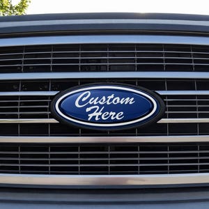 Könnte beinhalten: Nahaufnahme des Frontgrills eines Fahrzeugs mit einem individuellen ovalen Emblem. Das Emblem ist dunkelblau mit weißem Text, der "Custom Here" lautet. Der Grill hat horizontale Chromleisten.