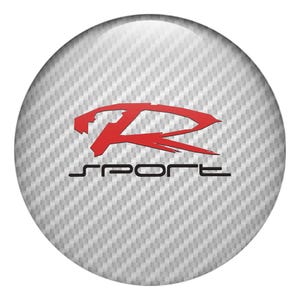 May include: A round emblem with a carbon fiber texture background. The emblem features the word "SPORT" in black, with a stylized red "R" logo above it. The emblem has a glossy finish.