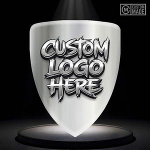Könnte beinhalten: Ein silbernes, schildförmiges Emblem mit dem Text "CUSTOM LOGO HERE" in einer metallischen, stilisierten Schriftart. Das Emblem befindet sich auf einer dunklen Oberfläche mit einem Scheinwerfereffekt. In der oberen rechten Ecke befindet sich ein "CUSTOM MADE"-Logo.