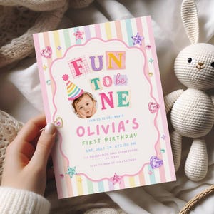 May include: A first birthday invitation card featuring pastel vertical stripes, a photo of a child in a party hat, and the text FUN TO BE ONE OLIVIA'S FIRST BIRTHDAY. The design includes pink scalloped borders, colorful stars, and heart accents.