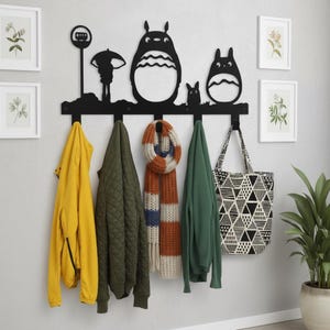 May include: Black metal wall-mounted coat rack featuring a silhouette design of characters from a popular animated film. The rack holds a yellow jacket, green quilted jacket, striped scarf, green sweater, and a tote bag with a geometric pattern.
