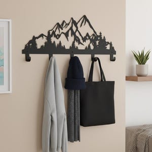 May include: Black metal wall-mounted coat rack with a mountain and forest silhouette design. The rack has four hooks and is holding a gray hoodie, a navy blue beanie and scarf, and a black tote bag. A houseplant sits on a nearby shelf.