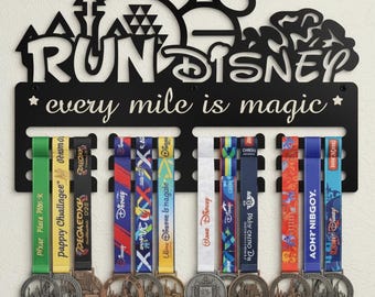 Può includere: Portamedaglie Run Disney in metallo nero con la scritta "every mile is magic". Il portamedaglie espone diverse medaglie di gara colorate. La parte superiore presenta una silhouette di Topolino e un design a castello. Le medaglie hanno vari colori e disegni di nastri.