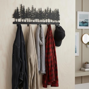 May include: A black metal wall-mounted coat rack featuring a forest silhouette design. Various garments, including a denim jacket, sweater, scarf, and plaid shirt, hang from the hooks. A black baseball cap is also displayed.