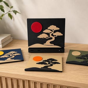 May include: A black rectangular electronic device with a red circle and a tan bonsai tree silhouette, accompanied by three square coasters featuring various color combinations of bonsai trees and suns in blue, tan, and olive green.