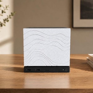 May include: A square electronic device featuring a textured white front panel with engraved horizontal wavy lines. The base is a solid black rectangular unit containing multiple USB ports, a small circular power button, and a tiny indicator light.