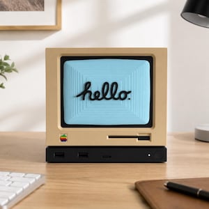 May include: A beige, retro-style computer monitor stand featuring a light blue screen with the word hello in black script. The front includes a small, multi-coloured apple logo and a black slot. It sits atop a black rectangular base with ports.