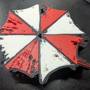 May include: A 3D-printed patch featuring the Umbrella Corporation logo. The design is a segmented umbrella shape with alternating red and white sections, outlined in black. The patch has a textured appearance.