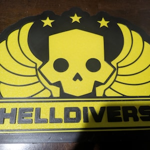 May include: A yellow and black emblem featuring a skull design with wings and three stars. The word "HELLDIVERS" is displayed in a yellow rectangular box at the bottom of the emblem. The emblem is likely a logo or patch.
