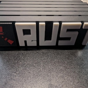 May include: A black and gray object with the word "RUST" in white block letters. The left side features a red and white logo. The object has a series of horizontal black bars on top.
