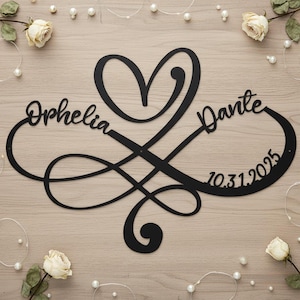 Op de afbeelding: Zwart metalen wanddecoratie met een hart en een oneindigheidssymbool. De namen "Ophelia" en "Dante" zijn gegraveerd, samen met de datum "10.31.2025". Een decoratief item, mogelijk voor een bruiloft of jubileum.