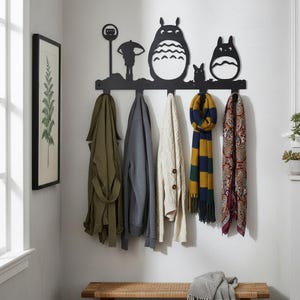 May include: Black metal wall-mounted coat rack featuring silhouettes of characters from a popular animated film. The rack holds several coats and scarves in various colors, including green, gray, and cream. A framed botanical print hangs on the wall.