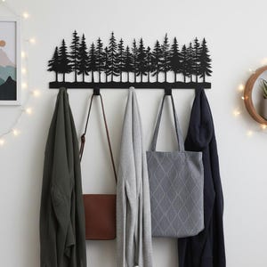 May include: A black metal wall-mounted coat rack featuring a silhouette of a forest. The rack has multiple hooks holding various items, including a green jacket, a brown purse, a gray sweater, a gray tote bag, and a navy blue jacket.