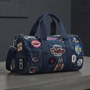 Peut inclure: Un sac de sport en jean bleu foncé doté de deux poignées supérieures et d'une bandoulière amovible. L'extérieur est orné de divers écussons brodés colorés, dont un logo Dallas bien visible, une bouche rose et plusieurs emblèmes circulaires.