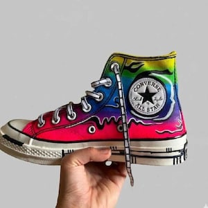 May include: A high-top sneaker featuring a vibrant rainbow gradient design with black drip-style outlines. The shoe includes a white circular Converse All Star patch on the side, white laces with black detailing, and a white rubber sole with black accents.