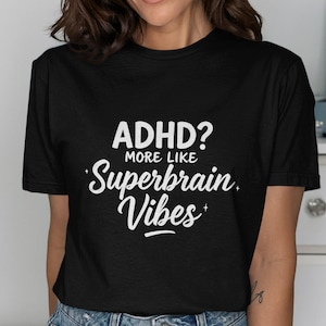 May include: Black t-shirt with the text "ADHD? More like Superbrain Vibes" in white script. The shirt is a crew neck and appears to be made of a soft material. The shirt is being worn.
