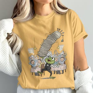May include: Mustard yellow t-shirt featuring a cartoon frog waiter balancing a tall stack of plates. The frog is in a tuxedo, and the background shows a restaurant scene with patrons. The design includes forks, spoons, and plates.