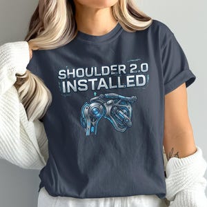 May include: Dark gray t-shirt with the text "SHOULDER 2.0 INSTALLED" above a blue and silver robotic shoulder illustration. The design has a futuristic, tech-inspired aesthetic.