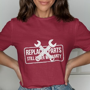 May include: Maroon t-shirt with a white graphic. The graphic features two crossed wrenches inside a white rectangle with the words "REPLACED PARTS STILL UNDER WARRANTY". The shirt is a crew neck and has short sleeves.