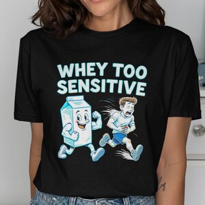 May include: Black t-shirt with the text "WHEY TOO SENSITIVE" above a cartoon illustration of a milk carton flexing and a person running while holding their stomach. The milk carton is smiling.