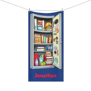 May include: A blue beach towel featuring a cartoon illustration of a locker filled with books, a baseball cap, and stickers. The locker door is open. The name "Jonathan" is printed in red at the bottom.