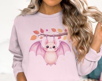 May include: A light pink sweatshirt featuring a cute cartoon bat hanging from a branch with autumn leaves. The bat has pink wings and a sweet expression. The branch has orange, yellow, and red leaves.