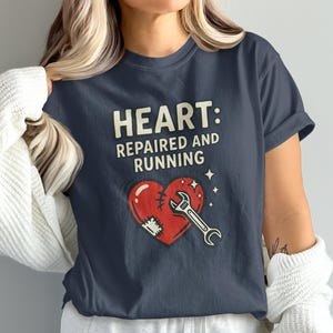May include: Navy blue t-shirt with the text "HEART: REPAIRED AND RUNNING". The graphic features a red heart with a wrench and a patch, with stars. The shirt is a casual wear item.