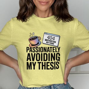 May include: A light yellow t-shirt with the text "PASSIONATELY AVOIDING MY THESIS". The shirt features a coffee cup graphic and a 404 error message. The shirt is a casual wear item.