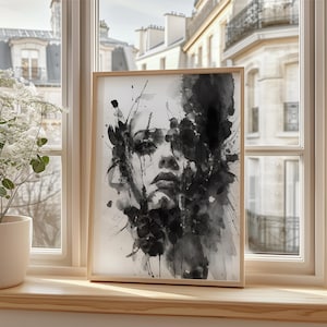 May include: A framed watercolour print of a woman's face in monochrome. The artwork features abstract brushstrokes and splatters, creating a dramatic portrait. The frame is light wood, and the artwork is displayed on a windowsill, with a view of buildings.