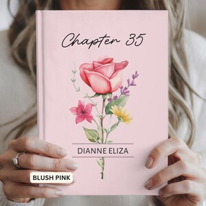 May include: A blush pink hardcover book featuring the text Chapter 35 in black script at the top. The center displays a watercolor illustration of a large pink rose with smaller pink and yellow flowers and green leaves, with the name DIANNE ELIZA below.