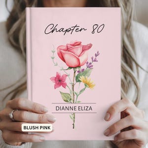 May include: A blush pink hardcover book featuring the text Chapter 80 in black script at the top. Below the text is a watercolor illustration of a large pink rose, a small pink flower, a yellow flower, and green leaves. The name DIANNE ELIZA is printed in black capital letters between two horizontal lines.