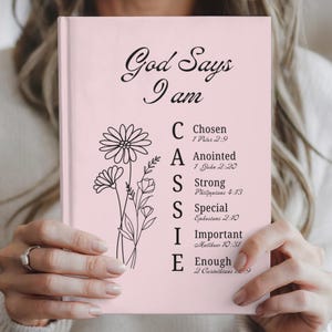 May include: A pink journal with the words "God Says I am" in black script. The journal features the name "CASSIE" in large letters, with each letter representing a positive affirmation. Floral line art decorates the left side of the journal.