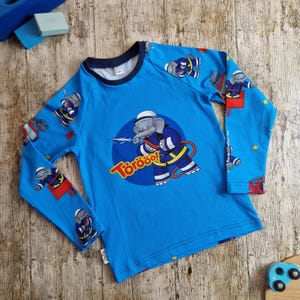 Könnte beinhalten: Ein blaues Langarmshirt mit einem Cartoon-Elefanten als Feuerwehrmann, der Wasser aus einem Schlauch spritzt. Das Shirt hat einen marineblauen Ausschnitt und das Wort "Törööö!" ist auf der Vorderseite aufgedruckt.