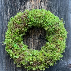 Könnte beinhalten: Ein kreisförmiger Kranz aus leuchtend grünem Moos, mit braunen Akzenten, vor einem dunklen, verwitterten Holzgrund. Die Textur des Kranzes ist dicht und natürlich, was auf einen handgefertigten Artikel für die Heimdekoration hindeutet.