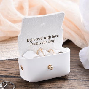 Könnte beinhalten: Eine weiße Ringbox aus Kunstleder mit goldenem Verschluss, die mehrere Ringe enthält. Auf der Innenseite des Deckels steht der Text "Delivered with love from your Boy". Die Box steht auf einer Holzoberfläche, mit einer weißen Blume und anderen dekorativen Elementen.