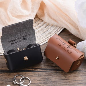 May include: Two leather ring boxes, one black and one brown, with gold hardware. The black box is open, revealing the text "Delivered with love from your Boy." The brown box has the text "v+h 18.12.2025." Two silver rings with gemstones are in the foreground.