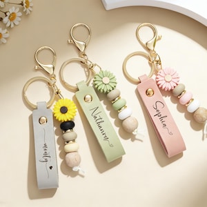 May include: Three personalised keyrings with gold-coloured hardware. Each keyring features a leather strap with a name, a flower charm, and wooden beads. The keyrings are in grey, green, and pink, with the names "monty", "Nathan", and "Sophia".