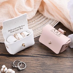 Puede incluir: Dos pequeñas cajas de anillos personalizadas de cuero blanco y rosa sobre una superficie de madera. La caja blanca está abierta, mostrando anillos y el texto "Delivered with love from your Boy". La caja rosa está cerrada, con las iniciales "A&E" y la fecha "18.12.2025".