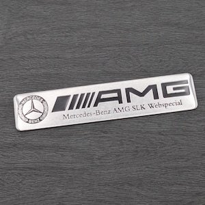 May include: A silver Mercedes-Benz AMG SLK Webspecial badge. The rectangular badge displays the Mercedes-Benz logo on the left, the AMG logo in the centre, and the text "Mercedes-Benz AMG SLK Webspecial" below. The badge is set against a dark textured background.
