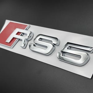 May include: Shiny chrome RS5 emblem with a red background on the letter R. The emblem is mounted on a white backing, with a dark grey background. The letters are in a bold, sans-serif font.