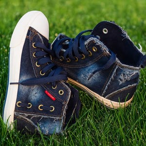 May include: Denim-style high-top trainers with white soles and navy blue laces. The shoes have a distressed appearance with brass eyelets and a small red tag. They are placed on green grass.