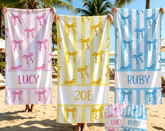May include: Three rectangular beach towels featuring vertical stripes and a repeating bow pattern. The left towel is pink and white with the name LUCY, the center is yellow and white with ZOE, and the right is blue and white with RUBY. Two matching drawstring bags are below.