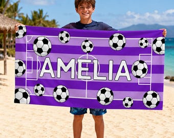 May include: A rectangular beach towel featuring a purple and light purple striped background with a white soccer field graphic. The towel displays the name AMELIA in large white capital letters, surrounded by multiple black and white soccer balls.