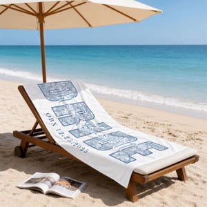May include: A white beach towel featuring the large blue text "BRIDE" with a floral pattern inside the letters. The text "OBX 12.25.2025" is printed in smaller blue font below the word. The towel is draped over a wooden lounge chair.