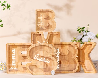 May include: A collection of wooden letter-shaped piggy banks with clear acrylic fronts. Each bank features a unique letter of the alphabet and is personalized with a white script name, including Emma, Sophia, and Brooklyn, displayed in a stack.